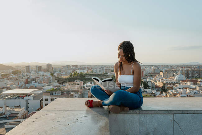 Trendige ethnische Frau in Sandalen liest Lehrbuch, während sie mit überkreuzten Beinen auf Zaun in der Stadt sitzt — Stockfoto