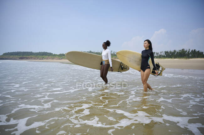 Smiling black sportswoman with longboard against Asian girlfriend with surfboard looking forward in ocean under cloudy blue sky — Stock Photo