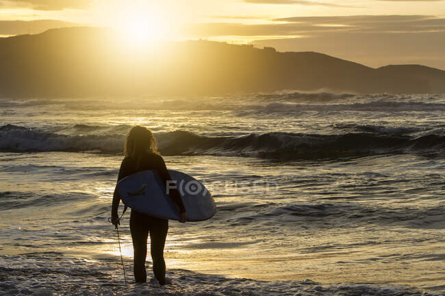 Visão traseira de uma jovem irreconhecível com prancha de surf entrando no mar durante o pôr do sol na praia em Astúrias, Espanha — Fotografia de Stock