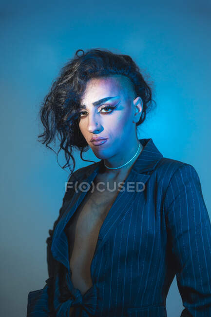 Young transsexual male model with makeup and tattoo in stylish jacket looking at camera on blue background — Stock Photo