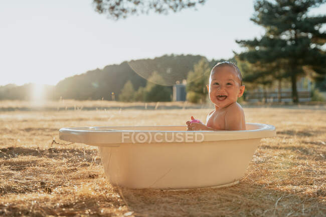 Вид сбоку счастливого малыша с игрушкой, сидящего в пластиковой ванне и играющего с водой в сельской местности — стоковое фото