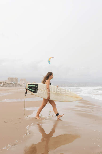 Vue latérale d'une jeune sportive heureuse en maillot de bain avec planche de surf regardant loin sur la côte sablonneuse contre l'océan orageux — Photo de stock