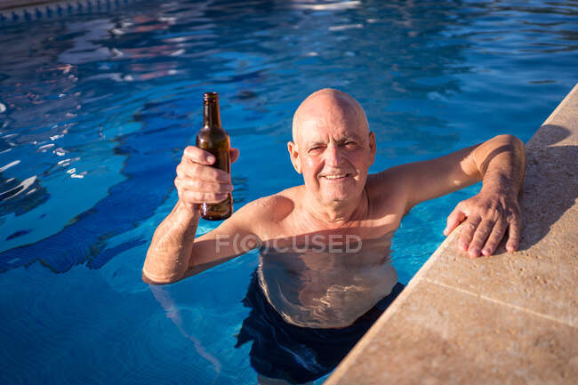 Angolo alto di rilassato anziano maschio bere birra da bottiglia di vetro mentre nuota in piscina — Foto stock