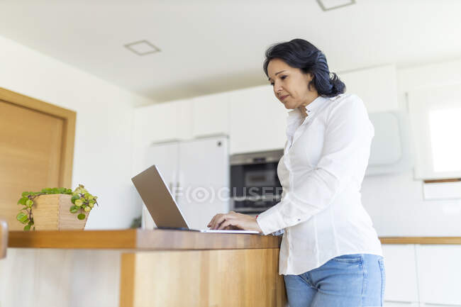 Focalizzata matura freelance navigando su Internet su netbook lavorando su un nuovo progetto mentre in piedi al piano di lavoro in cucina a casa — Foto stock