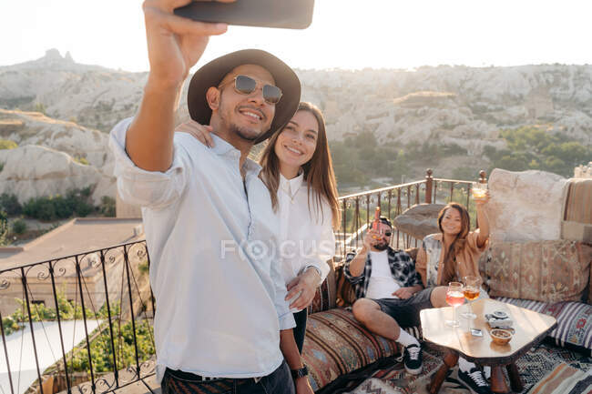 Grupo de jóvenes y diversos amigos tomando selfie en el teléfono celular mientras beben cócteles en el bar de la terraza en Capadocia, Turquía - foto de stock