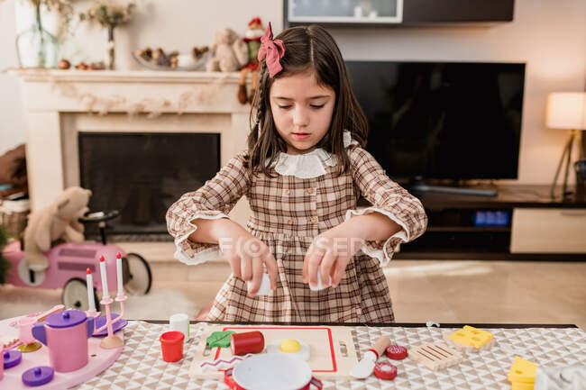 Aufmerksames Kind im karierten Kleid spielt mit Spielzeug, während es beim Kochen am Tisch im hellen Raum Eier bricht — Stockfoto