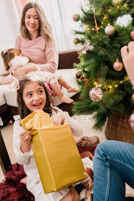 Sourire enfant ouverture boîte cadeau entre le père de la culture et la mère allaitement bébé pendant les vacances du Nouvel An à la maison — Photo de stock