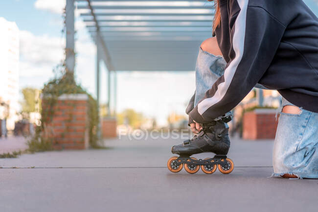 Vue latérale d'une jeune femelle non reconnaissable portant un pull noir et un jean bleu clair avec des fentes boutonnant des lames de rouleau debout sur un genou dans la rue — Photo de stock