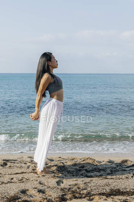 Vue latérale de la femelle ethnique penchée en arrière tout en se tenant à Ashta Chandrasana pose pendant la pratique du yoga sur la côte sablonneuse contre l'océan — Photo de stock