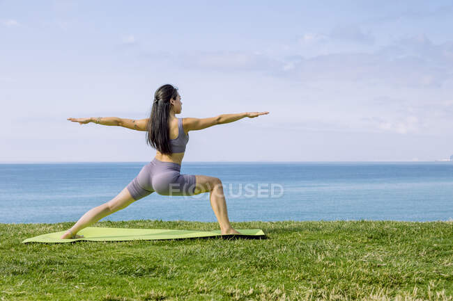 Vista posterior de la hembra étnica descalza que realiza la pose Virabhadrasana en la esterilla de yoga contra el océano sin fin bajo el cielo nublado - foto de stock
