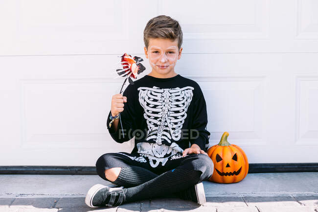 Повне тіло моторошного хлопчика в чорному костюмі скелета з макіяжем, що тримає квітку і дивиться на камеру, сидячи біля гарбузового ліхтаря на білій стіні — стокове фото