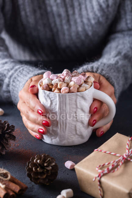 From above of crop female holding mug of hot drink with marshmallows among Christmas gifts cones and cinnamon sticks — Stock Photo