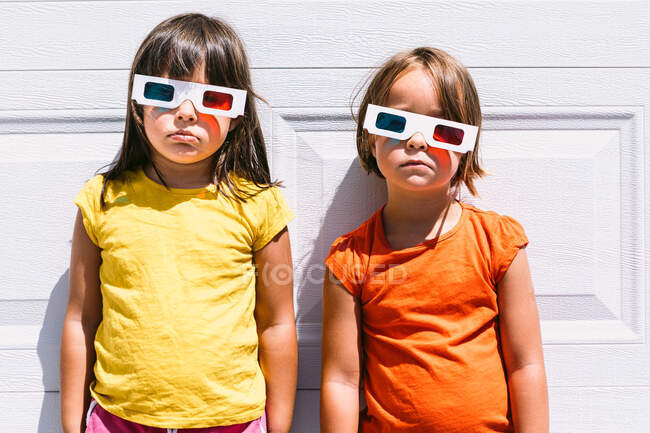 Милі дівчата в повсякденному барвистому одязі і тривимірних окулярах, що стоять на білому тлі стіни, дивлячись на камеру — стокове фото