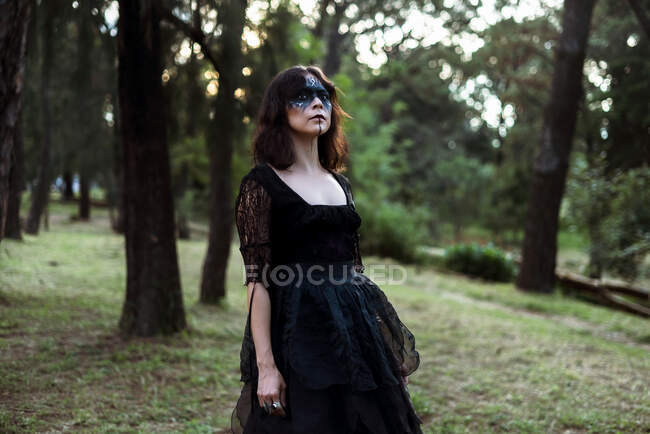 Mystic strega in abito lungo nero e con viso dipinto in piedi guardando lontano in oscuri boschi cupi — Foto stock