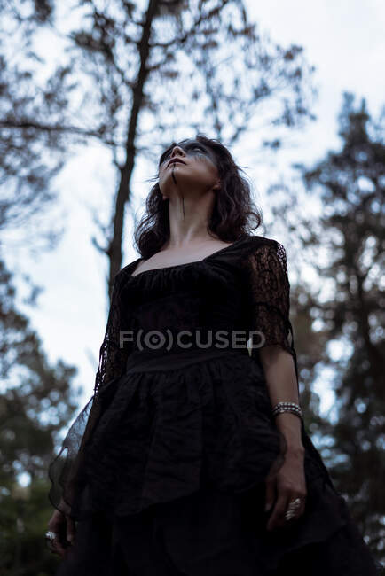 Von unten eine mystische Hexe im langen schwarzen Kleid und mit geschminktem Gesicht, die in dunklen, düsteren Wäldern wegschaut — Stockfoto