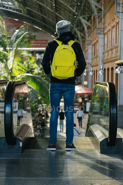 Vista trasera de hipster masculino anónimo con mochila de color amarillo brillante de pie cerca de escaleras mecánicas en el gran centro comercial - foto de stock
