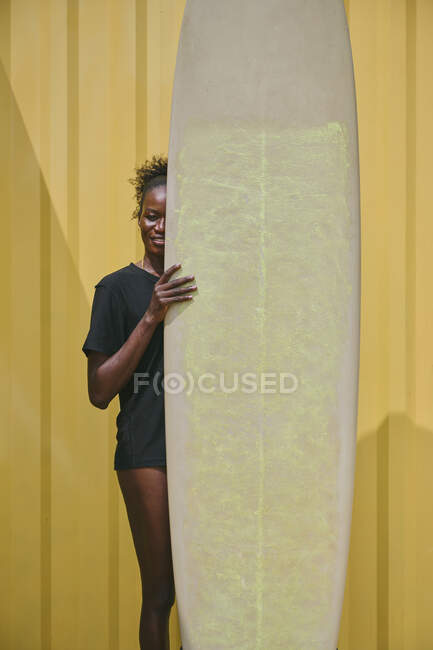 Cheerful young African American sportswoman in bikini and t shirt looking at camera with surfboard in a yellow container on the coast — Stock Photo