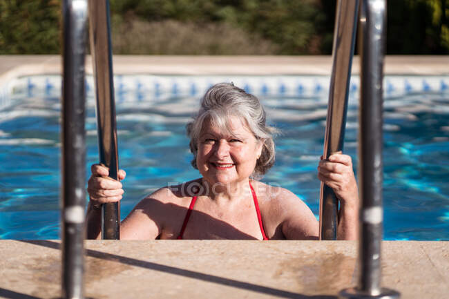 Mulher sênior positiva em roupa de banho descendo na piscina e segurando corrimãos inoxidáveis enquanto relaxa no dia ensolarado — Fotografia de Stock