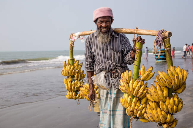 INDIA, BANGLADESH - DECEMBER 4, 2015: Ethnic male farmer in casual clothes walking on the beach carrying bunch of fresh bananas looking at camera — Stock Photo