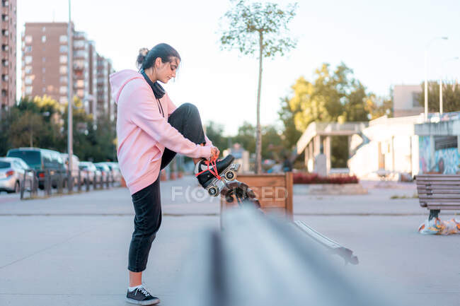 Vue latérale du corps entier positif jeune femme portant un sweat à capuche rose clair et un pantalon noir lacant les patins à roulettes s'appuyant sur la jambe à l'arrière du banc en bois en partie — Photo de stock