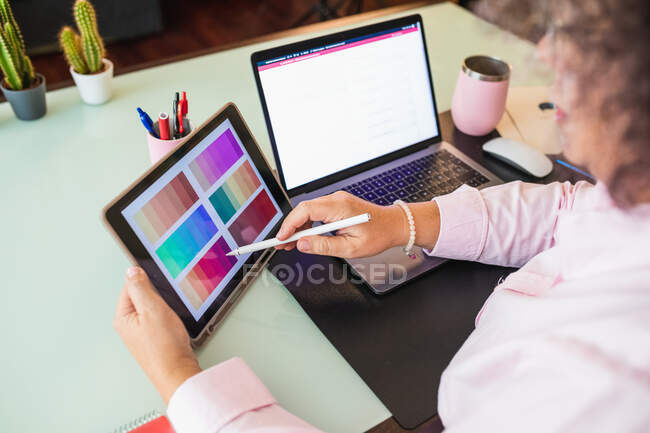 Crop senior female touching screen on tablet while pointing at color palette and speaking during video chat on netbook in office — Stock Photo