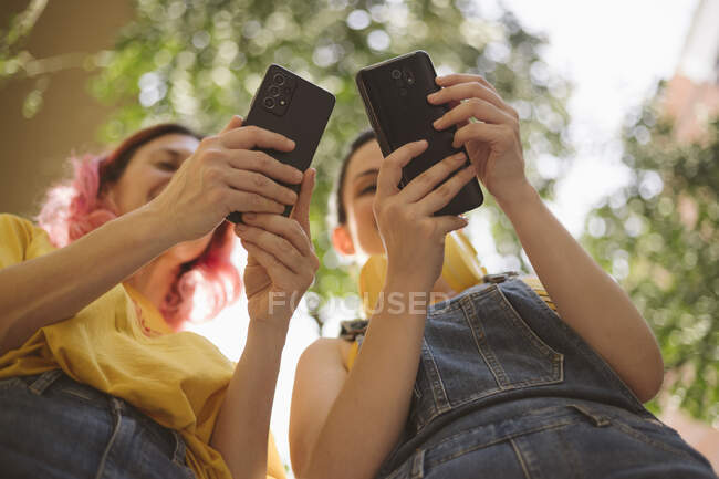 From below of positive female friends in casual clothes standing close and browsing mobile phones together — Stock Photo