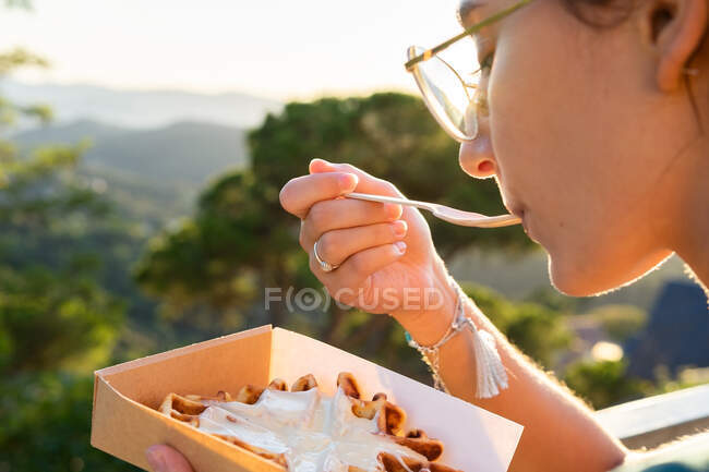 Seitenansicht der Ernte junge Frau essen leckere belgische Waffeln mit Schlagsahne in Take-away-Box gegen Berge im hinteren beleuchteten — Stockfoto