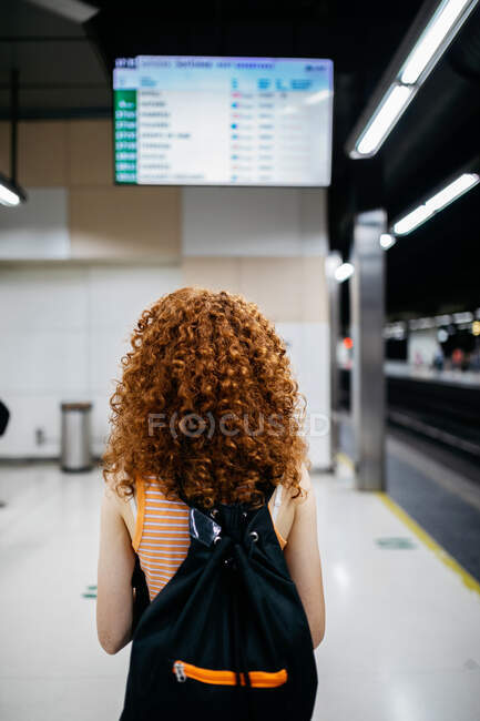 Back view of anonymous female tourist with backpack and curly hair against arrival display in underground station — Stock Photo
