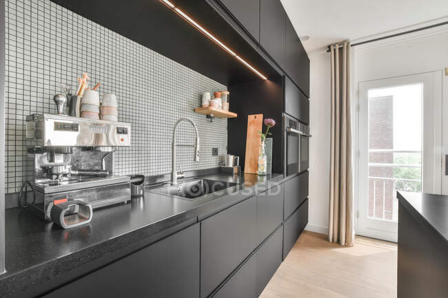 Máquina de café moderna colocada no balcão da cozinha escura perto da pia na cozinha moderna com armários pretos no apartamento durante o dia — Fotografia de Stock
