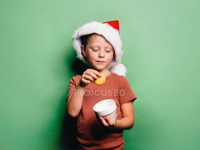 Menino adorável com chapéu de Natal Papai Noel tomando biscoito de xícara contra fundo verde — Fotografia de Stock