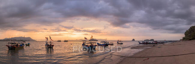 Ângulo largo de barcos de pesca com bandeiras nacionais ondulando na costa arenosa molhada lavada pelo mar sob o céu nublado sombrio no por do sol em Malaysia — Fotografia de Stock