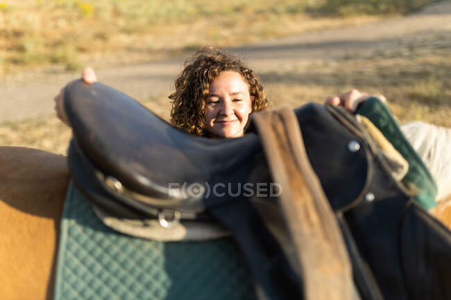 Middle aged female with curly hair putting saddle on back of mare on farm in sunlight — Stock Photo