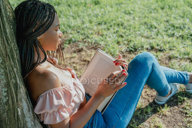Vista lateral de la joven y soñadora hembra negra con libro de texto y trenzas afro mientras está sentada en el césped - foto de stock