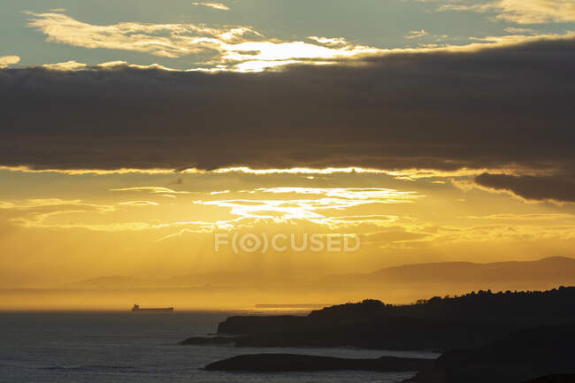Picturesque scenery of hilly seacoast and sundown sky with sun rays penetrating clouds over calm sea with distant vessel in El Musel seaport in Asturias Spain — Stock Photo