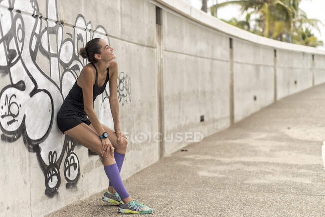 Side view of positive athletic female runner in sportswear leaning on wall while taking break during workout in city and looking up — Stock Photo