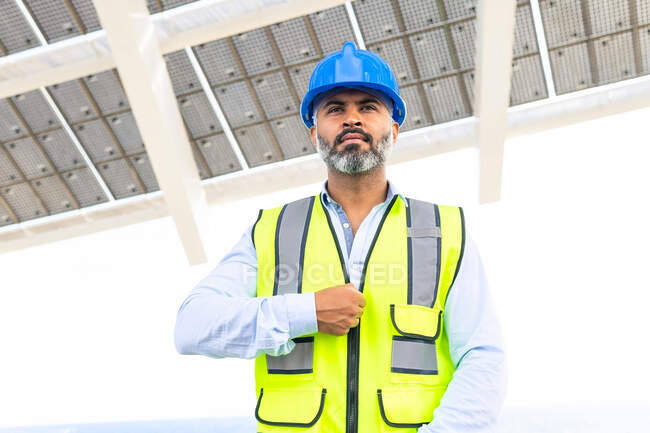 Entrepreneur masculin ethnique mature en casque et gilet debout regardant loin près de la centrale solaire — Photo de stock