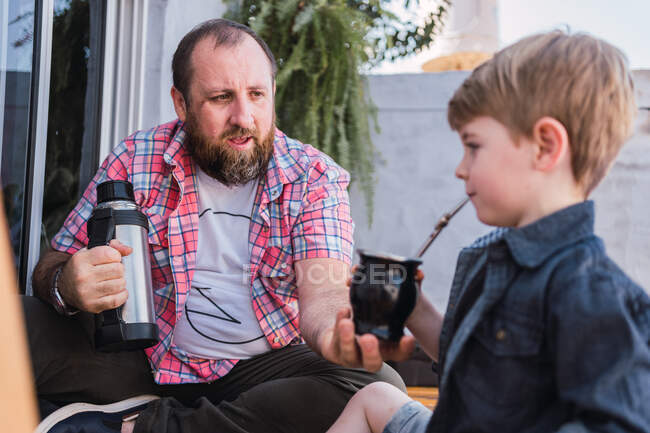 Sincere child with calabash gourd of infused drink against cheerful bearded dad with thermos on blurred background — Stock Photo