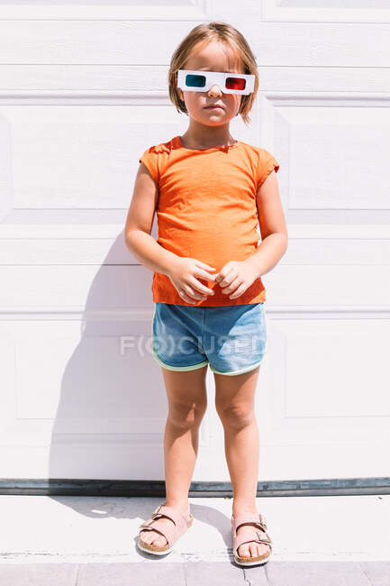 Linda chica en ropa colorida casual y gafas tridimensionales de pie sobre fondo de pared blanca mirando a la cámara - foto de stock