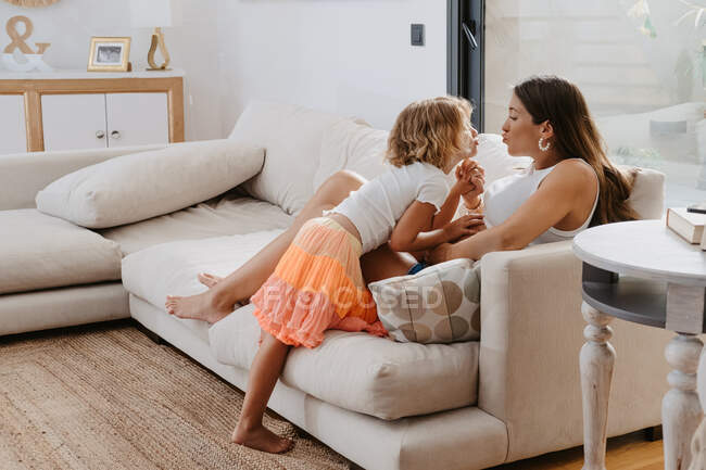 Visão lateral da mulher e da menina expectante com lábios amuados de mãos dadas enquanto olham um para o outro no sofá em casa — Fotografia de Stock