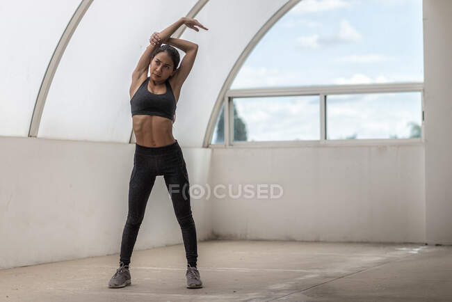 Atleta joven de etnia femenina en uso activo con los brazos levantados mirando hacia adelante durante el entrenamiento de estiramiento durante el día - foto de stock