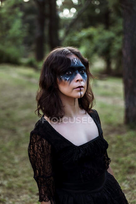 Mystic witch in long black dress and with painted face standing looking away in dark gloomy woods — Stock Photo
