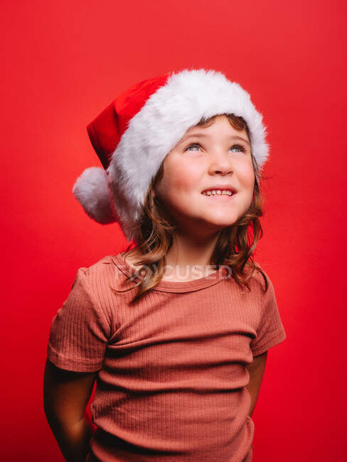 Adorável menina feliz em roupas casuais e chapéu de Papai Noel sorrindo enquanto em pé contra o fundo vermelho e olhando para longe — Fotografia de Stock