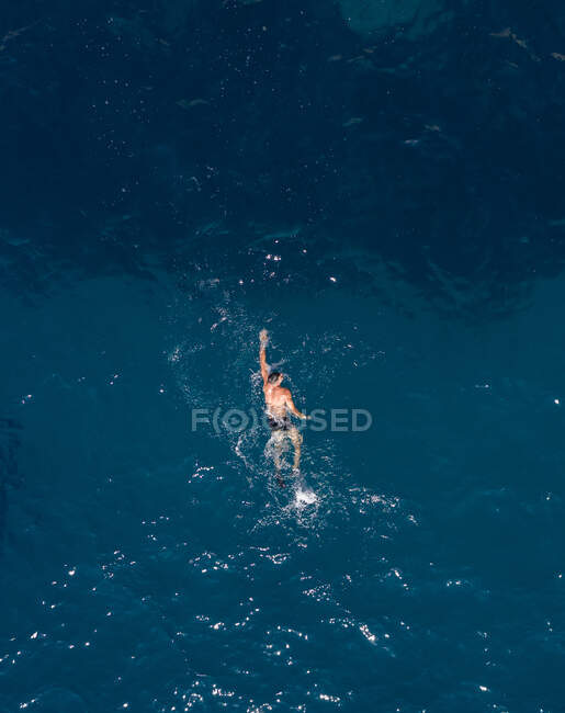 Drone vista de turista masculino anónimo con torso desnudo nadando en el océano ondulado durante el viaje a la luz del día - foto de stock