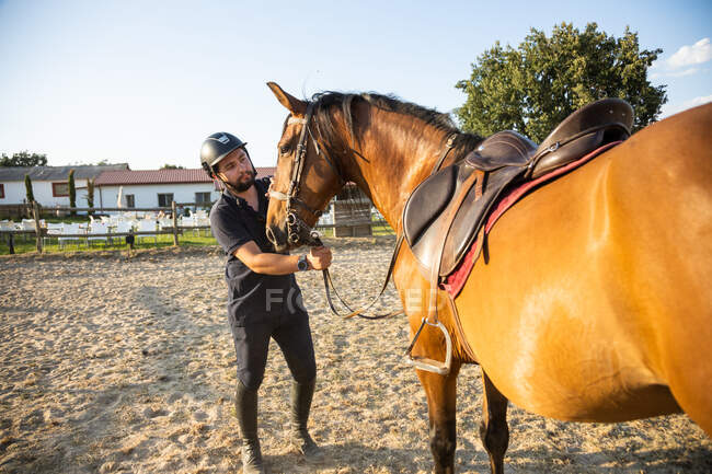 Adult male in protective helmet holding stallion by reins against stables of riding school in countryside — Stock Photo