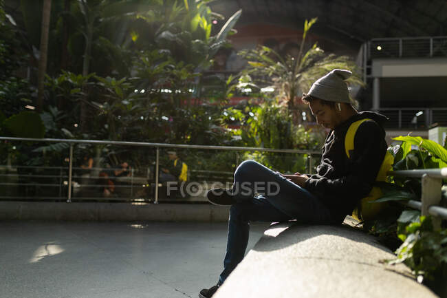 Hipster afroamericano maschio con zaino seduto sul bordo di pietra in strada e messaggistica sul cellulare e ascoltare musica su auricolari — Foto stock