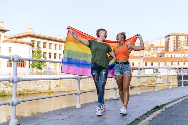 Mulher tatuada legal com mohawk e bandeira LGBTQ abraçando namorada com olhos fechados contra o canal na cidade — Fotografia de Stock