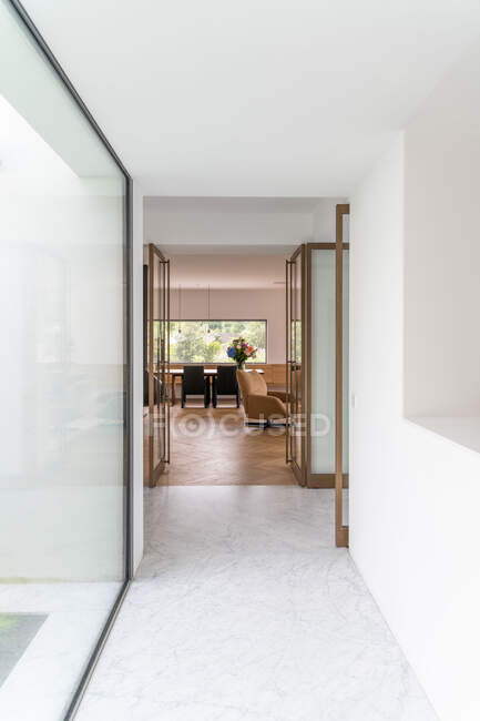 Estrecho pasillo con paredes de vidrio que conduce a una amplia sala de estar con cómodo sillón y comedor en villa contemporánea a la luz del día - foto de stock