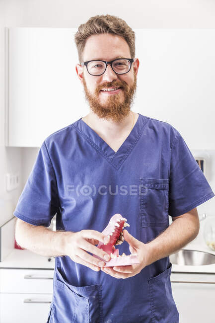 Позитивний бородатий чоловік у формі та окулярах зі штучною щелепою тварин у ветеринарній клініці та дивлячись на камеру — стокове фото