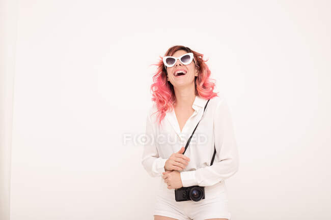 Mujer feliz con pelo rosa en gafas de sol y ropa elegante de pie con cámara fotográfica sobre fondo blanco en la habitación - foto de stock
