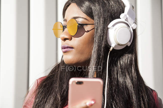 Нахабна молода афроамериканка з довгим волоссям у червоній куртці та сонцезахисних окулярах слухає музику через навушники та мобільний телефон, насолоджуючись сонячним днем у місті. — стокове фото